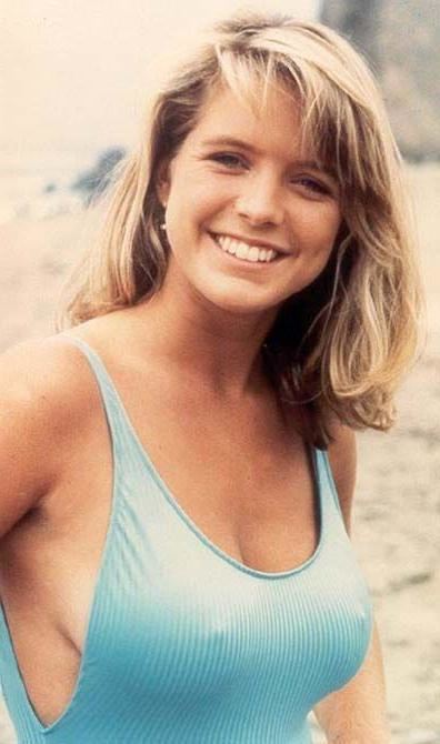 Courtney Thorne-Smith smiling, an American actress with medium-length wavy blonde hair, has earrings, braless, wearing a light blue low-round neck one-piece with visible cleavage.