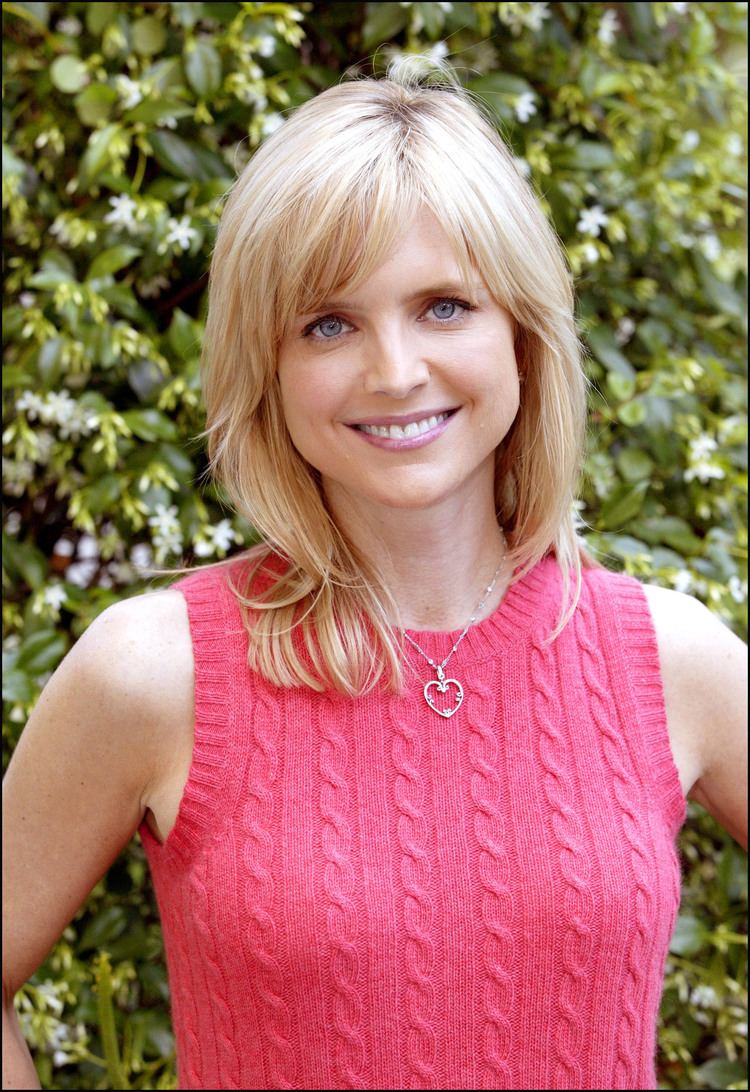 Courtney Thorne-Smith smiling with hands on her hips, an American actress with medium-length blonde hair with bangs, has light gray eyes, a silver heart-shaped pendant necklace, wearing a pink knitted-sleeveless top.
