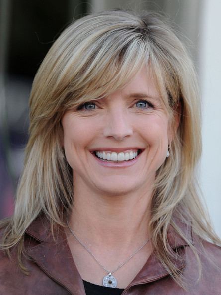 Courtney Thorne-Smith smiling, an American actress with a blonde layered haircut styled straight with side-swept bangs at the John Varvatos 7th Annual Stuart House Benefit in L.A. on March 8, 2009. She has light-gray eyes, wearing a  pair of pearl earrings, a silver pendant necklace, a black inner blouse, and a chocolate-brown coat.