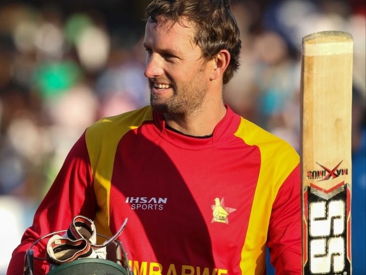 Craig Ervine is smiling while looking to his right, has brown hair, a beard, and a mustache, and his right hand holds a black helmet with a red-white-black chin strap, on his left is a Cricket Bat with an SS print, he is wearing his red-yellow National Cricket uniform with a print on his right chest “JHSAN SPORTS” at the center is “ZIMBABWE” and on left is a yellow print.