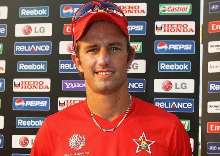 Craig Ervine is smiling, and has brown hair, a shaved beard, and a mustache, at the back are different kinds of brands, he is wearing a red cap and sunglasses, a silver necklace, and a red shirt with a star print on his left chest, and a ball on his right chest with the  “Cricket World Cup”.