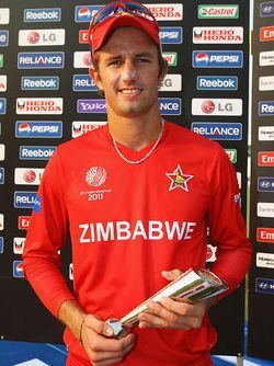 Craig Ervine is smiling, has brown hair, a shaved beard, and a mustache, both hands holding a trophy, at the back are different kinds of brands, he is wearing a red hat and sunglasses, a silver necklace, and a red long-sleeve shirt with the print “ZIMBABWE” on the center and a star on his left chest, and a ball with the year 2011 on right.