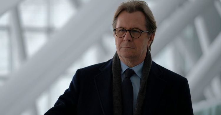 Gary Oldman looking afar while wearing a black coat, blue long sleeves, black necktie, and eyeglasses in a scene from the 2021 film, Crisis