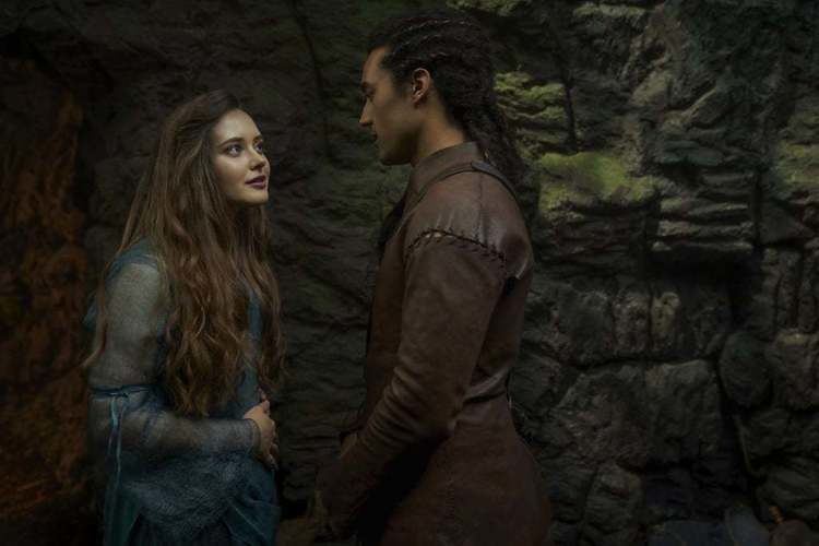 Katherine Langford as Nimue smiling while looking at Devon Terrell as Arthur in a scene from the 2020 American TV series Cursed (2020). Katherine having long curly hair and wearing a blue dress while Devon wearing a brown coat.
