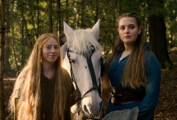Katherine Langford as Nimue with a serious face while Lily Newmark as Pym smiling and standing beside a white horse and trees in the background in a scene from the 2020 TV series Cursed. Katherine has long curly hair and wearing a green dress while Lily is having long blonde hair and wearing a brown dress