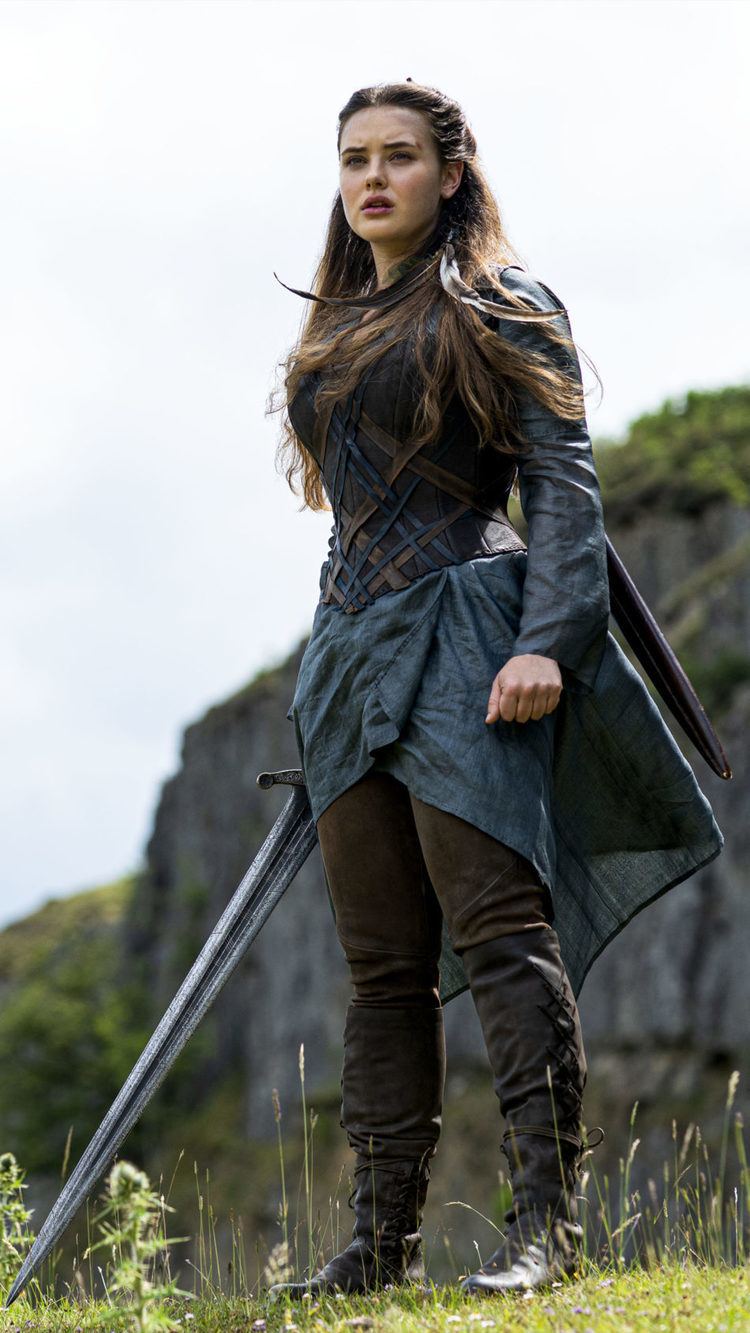Katherine Langford as Nimue looking serious with long hair and holding a sword in a scene from the 2020 American series "CURSED" with a mountain in the background and wearing a dress, brown pants, and black boots