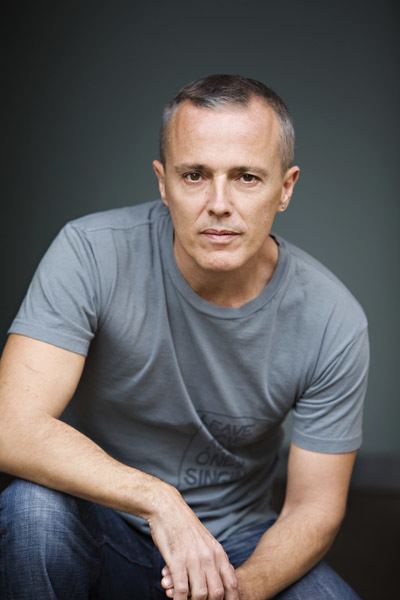Curt Smith's arms leaning on his knee and leg with a serious look while wearing a gray t-shirt and denim pants.