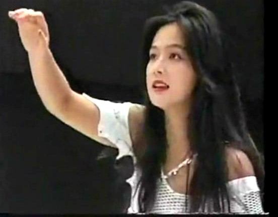 Cutie Suzuki smiling while looking at her hands, with long black hair, and wearing a white off-shoulder top.