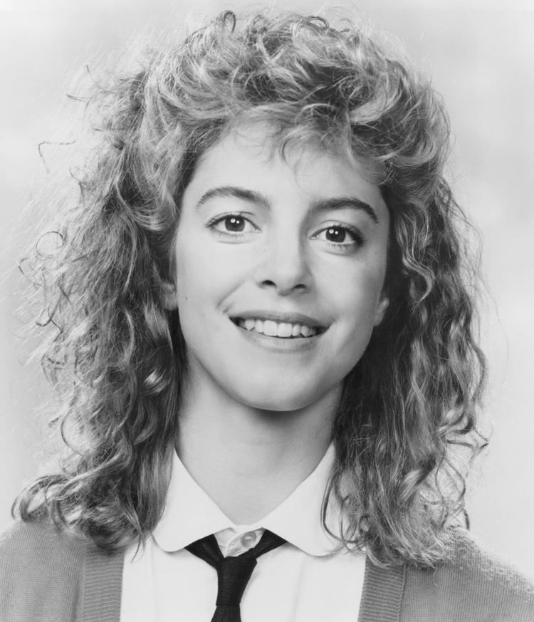 Cynthia Gibb smiling with curly hair and wearing a white shirt, a black necktie and knitted cardigan