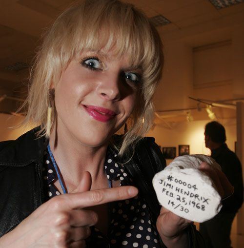 Cynthia Plaster Caster with a creepy smile while pointing a white stone with Jimi Hendrix's name, with blonde hair, wearing earrings, a black jacket, and a white and black polka dot top.