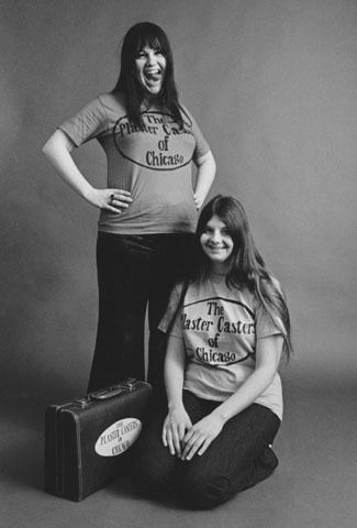 Cynthia and Dianne Plaster Caster are smiling with a black suitcase on the floor. Cynthia standing, wearing a shirt and black pants while Dianne sitting on the floor, wearing a shirt and black pants.