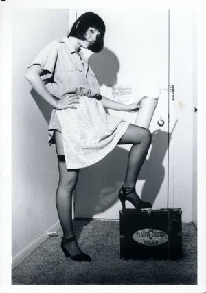 Cynthia Plaster Caster's side-view pose with her hand on her waist while stepping a box and holding a jar, with short black hair, wearing a polo shirt, white skirt, and black shoes.