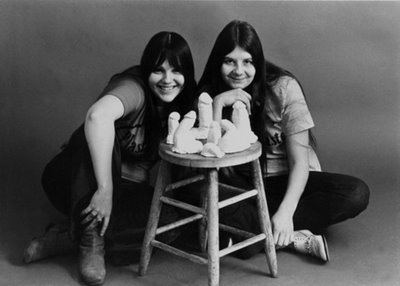 Cynthia and Dianne Plaster Caster are smiling while sitting on the floor with a chair full of plaster casts of persons' erect penises. Both are wearing shirts and black pants.