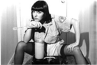 Cynthia Plaster Caster with a serious face while sitting on a chair while her hands are on her chin and thigh, with short black hair, wearing a polo shirt, and white skirt.