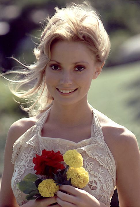 Cynthia Wood smiling with her hair windswept and holding red and yellow flowers while holding a white sleeveless gown.