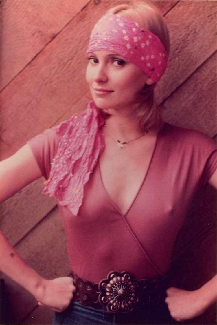 Cynthia Wood smiling with her hands in her waist wearing a pink bandana in her head and a v-neck shirt as well as jean pants.
