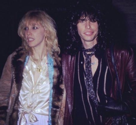 Cyrinda Foxe looking afar while wearing brown jacket and white inner blouse while Steven Tyler wearing brown leather jacket and striped long sleeves and neck tie