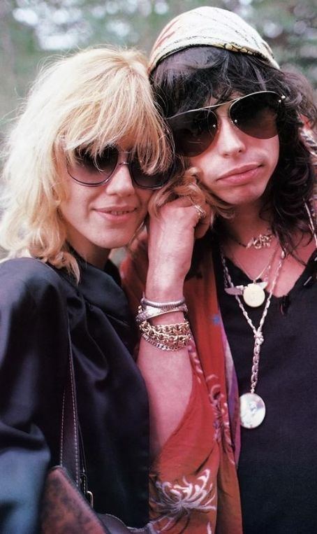 Cyrinda Foxe smiling and wearing shades and black coat while Steven Tyler wearing shades, bandana, red blazer and black inner shirt