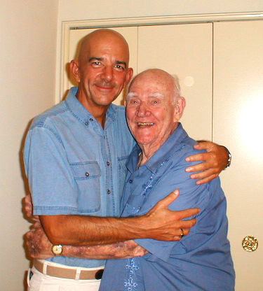 Jim Nolt and Dabbs Greer are smiling while hugging each other. Jim is wearing a light blue polo, beige belt, white pants, and wristwatch while Dabbs is wearing a blue polo and wristwatch