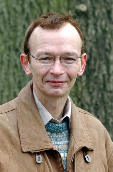 Dai Bradley smiling and wearing a white shirt under a blue knitted vest, brown jacket and eyeglasses