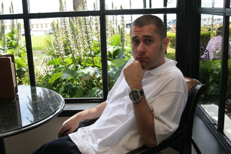 Damizza looking serious while sitting on a chair with his left hand on his chin with plants in the background, and wearing a white collared shirt, watch and pants