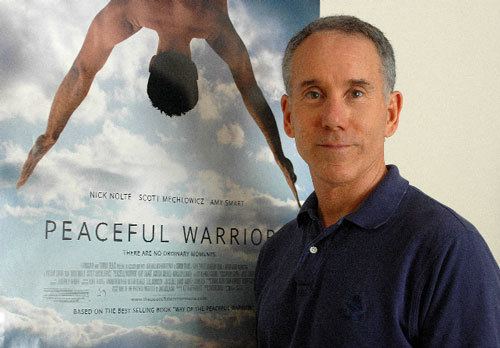 Dan Millman smiling and wearing a blue polo shirt beside the poster of the 2006 movie, Peaceful Warrior