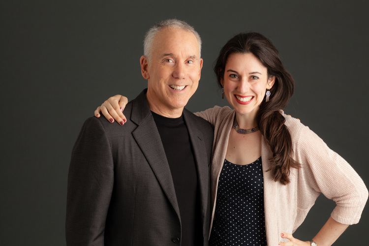 Dan Millman smiling and wearing a black coat and black shirt while  Sierra Prasada wearing a beige blazer and polka dot inner top