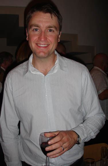 Daniel Casey smiling and holding a glass of wine while wearing white long sleeves