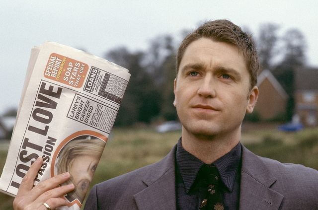 Daniel Casey looking afar while holding a newspaper and wearing a gray coat and black long sleeves