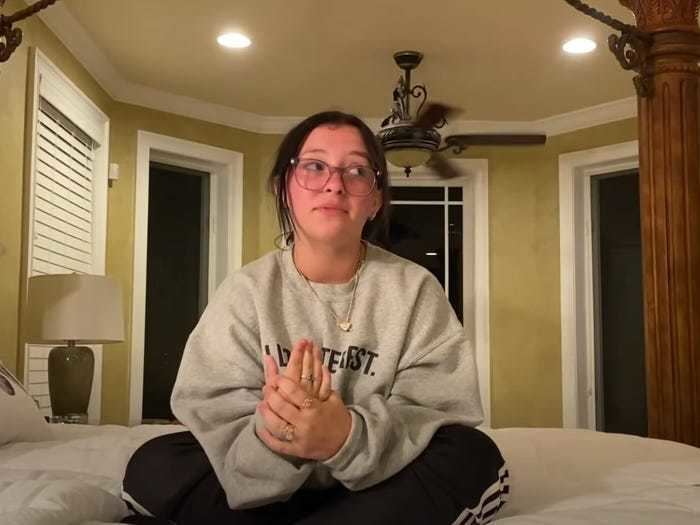 Danielle Cohn looking on her left side while sitting on a bed, wearing eyeglasses, a necklace, a gray sweatshirt, and a black with white striped pants.