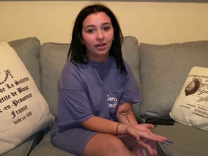 Danielle Cohn with mouth opens while sitting on a gray couch, with a tattoo on her arm, and wearing a purple shirt.