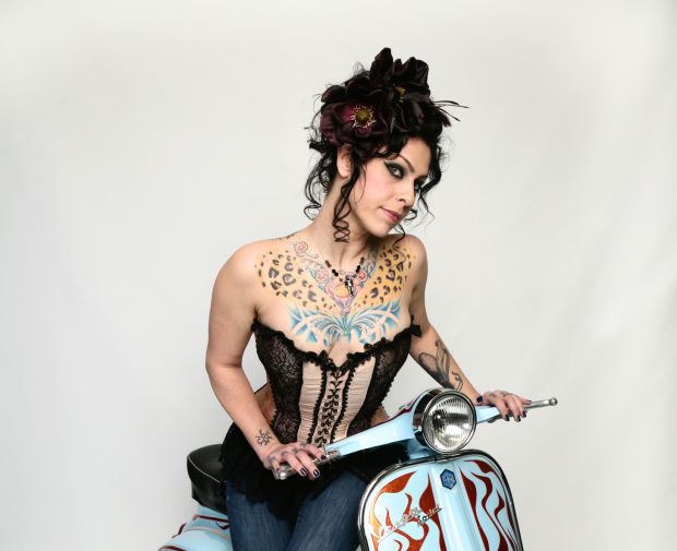 Danielle Colby with a tight-lipped smile while riding a scooter, with curly hair, with tattoo on her body, wearing a black tube top, and blue pants.