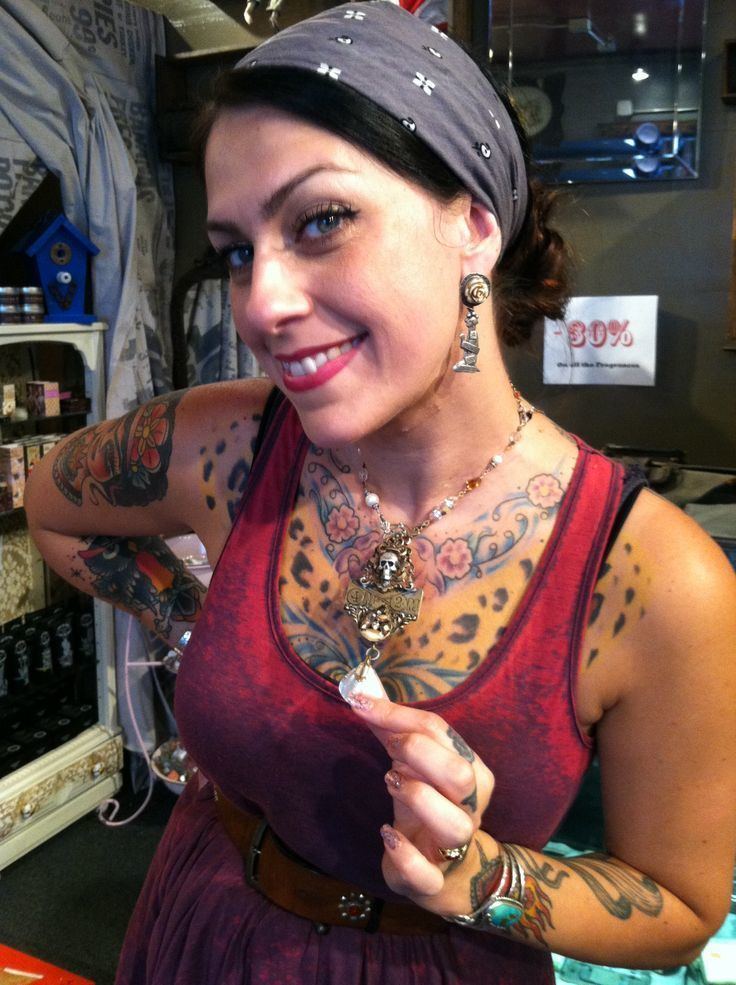 Danielle Colby smiling while holding her necklace, with a tattoo on her body, wearing a gray headband, earrings, and a red sleeveless dress with a brown belt.