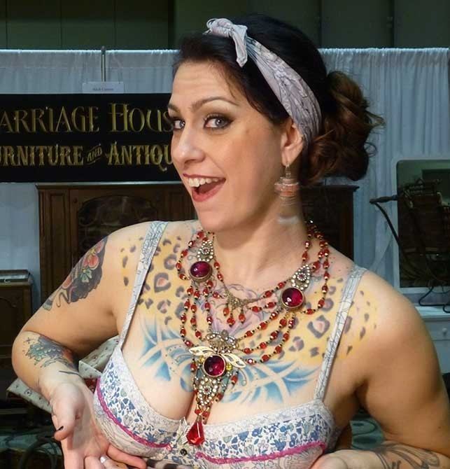 Danielle Colby smiling while holding her boobs, with a tattoo on her body, wearing a headband, a red necklace, and a multi-colored brassiere.