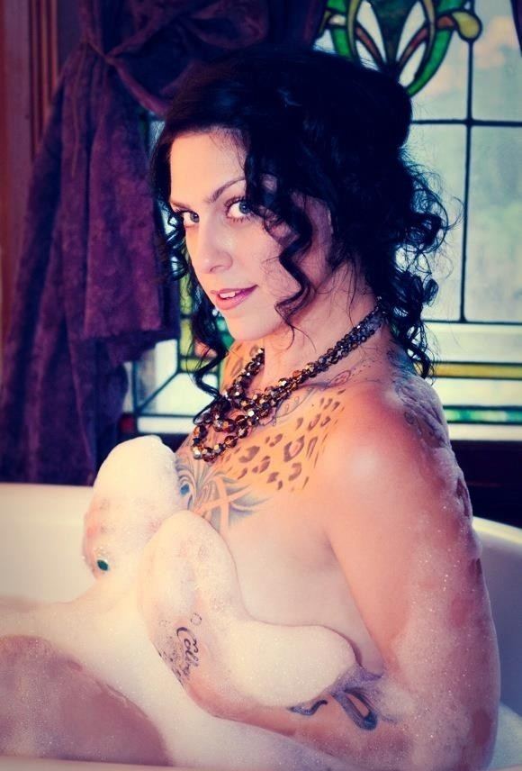 Danielle Colby smiling and naked while covering her boobs in a bathtub, with curly hair, with a tattoo on her body, and wearing a necklace.