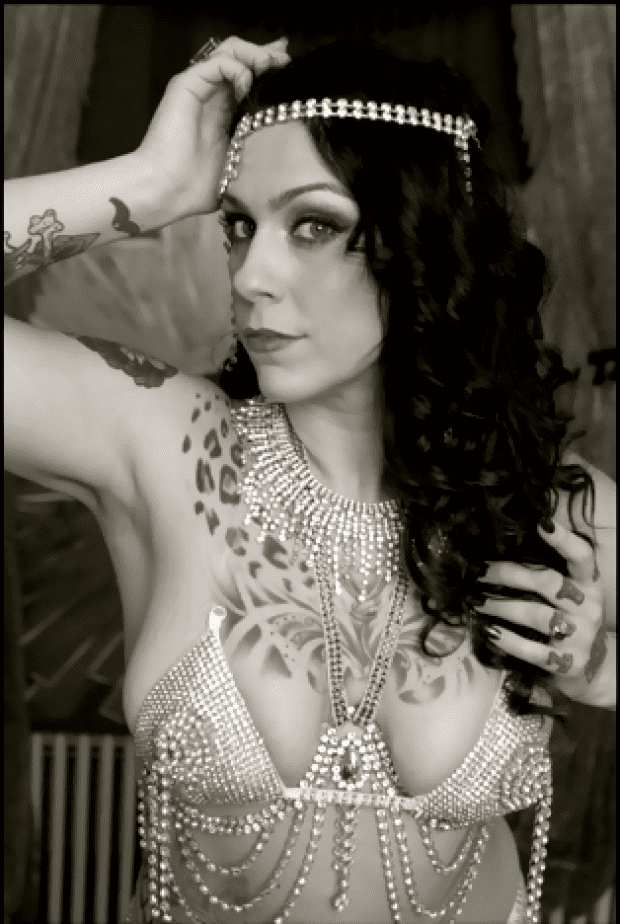 Danielle Colby with a serious face while holding her head, with a tattoo on her body, wearing a head chain, a necklace, and a fancy top.