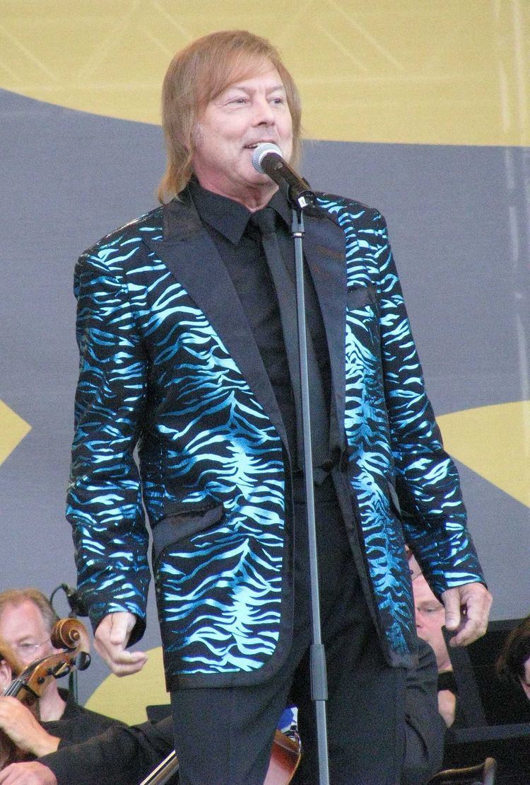 Danny (Finnish singer) singing, with blonde hair, wearing a black and blue suit, black long sleeves, and a tie.