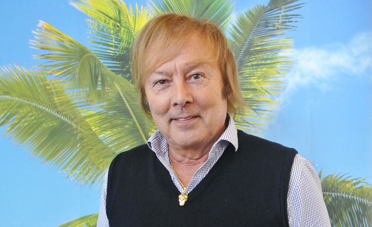 Danny (Finnish singer) smiling, with blonde hair and wearing a necklace and a black vest.