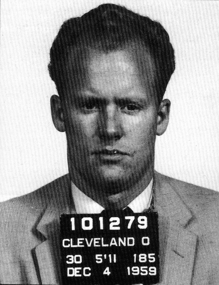 Danny Greene with a serious face and wearing a suit and a tie with a placard.