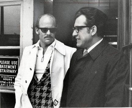 Danny Greene wearing eyeglasses, a black suit, and a tie together with his friend with a mustache, wearing sunglasses, and a white coat.