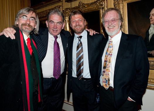 John Tams, Daragh O'Malley, Sean Bean, and Bernard Cornwell smiling together while wearing a black coat, white long sleeves, and necktie