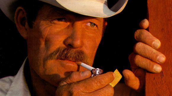 Darrell Winfield is serious, lighting up the cigarette while holding it with his mouth using a gold plated lighter in his right hand and left hand on the wall, has a mustache and brown hair wearing a white cowboy hat and a white polo.