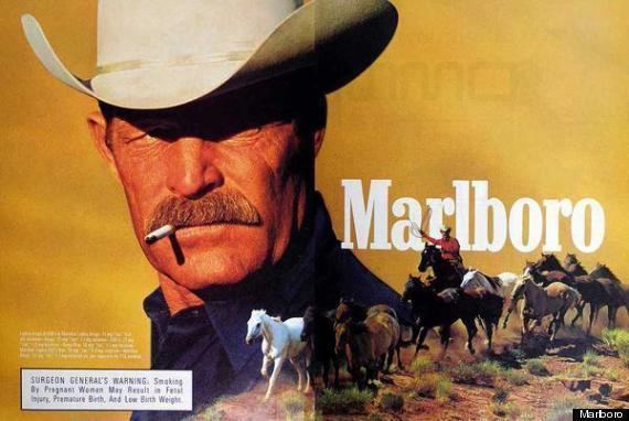 In a Marlboro advertisement, Darrell Winfield with a fierce look, looking at the right side while smoking, has brown hair and a mustache, wearing dark blue polo and a white cowboy hat. On the right a cowboy riding a horse along with eleven horses with his right hand up holding a rope, wearing white cowboy hat and red long sleeve polo.