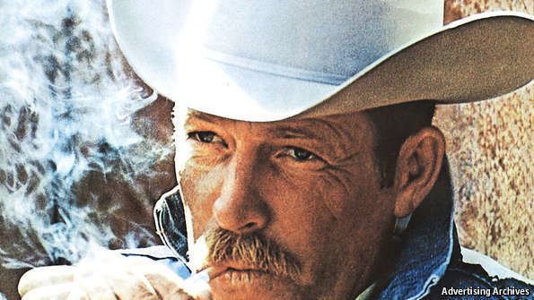 Darrell Winfield is serious, lighting up his cigarette while covering it with his left hand, has a mustache and brown hair wearing white cowboy hat and a denim jacket over a white polo.