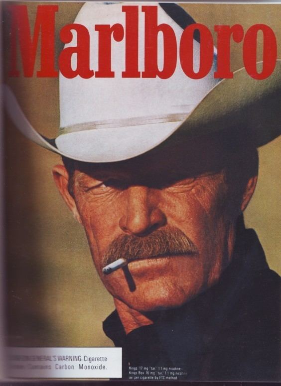 In a Marlboro advertisement, Darrell Winfield with a fierce look, looking at the right side while smoking, has brown hair and a mustache, wearing dark blue polo and a white cowboy hat.