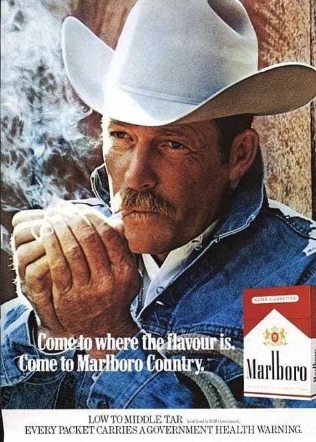 In a Marlboro advertisement Darrell Winfield is serious, lighting up his cigarette with his right hand while covering it with his left hand, has a mustache and brown hair wearing white cowboy hat with white rope on his arm and a denim jacket over a white polo.
