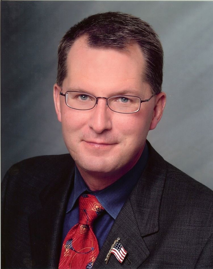 Dave Pelzer with a tight-lipped smile while wearing eyeglasses and blue long sleeve under a red necktie and black coat with American flag lapel pin