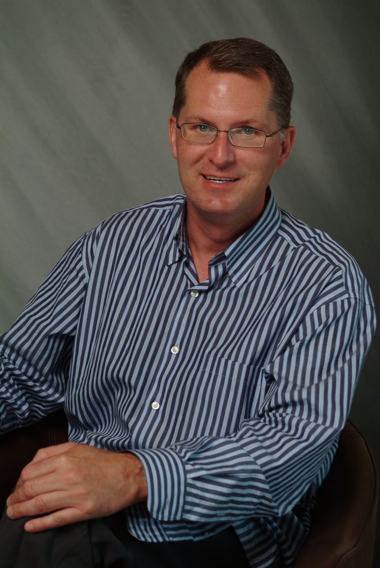 Dave Pelzer smiling and sitting on a chair with hand on his knees while wearing a white and blue striped long sleeve, black pants, and eyeglasses