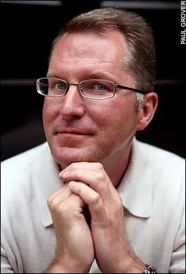 Dave Pelzer with a tight-lipped smile while his hands are together and wearing eyeglasses, a white polo shirt, and a wristwatch