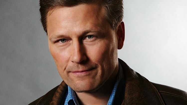 David Baldacci with a tight-lipped smile while wearing a brown jacket and blue checkered long sleeves